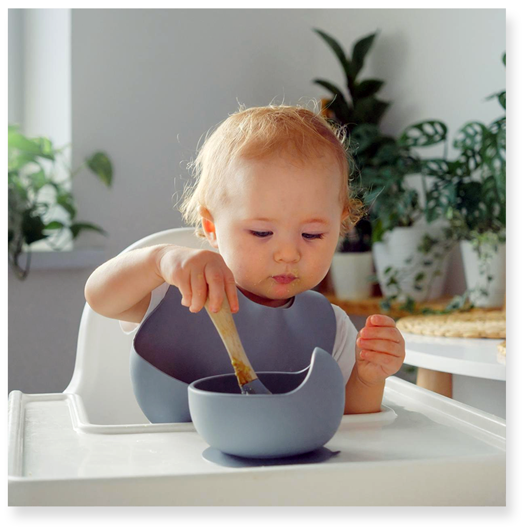 A young child sits in a high chair, stirring food in a gray bowl with a wooden spoon, surrounded by green plants.